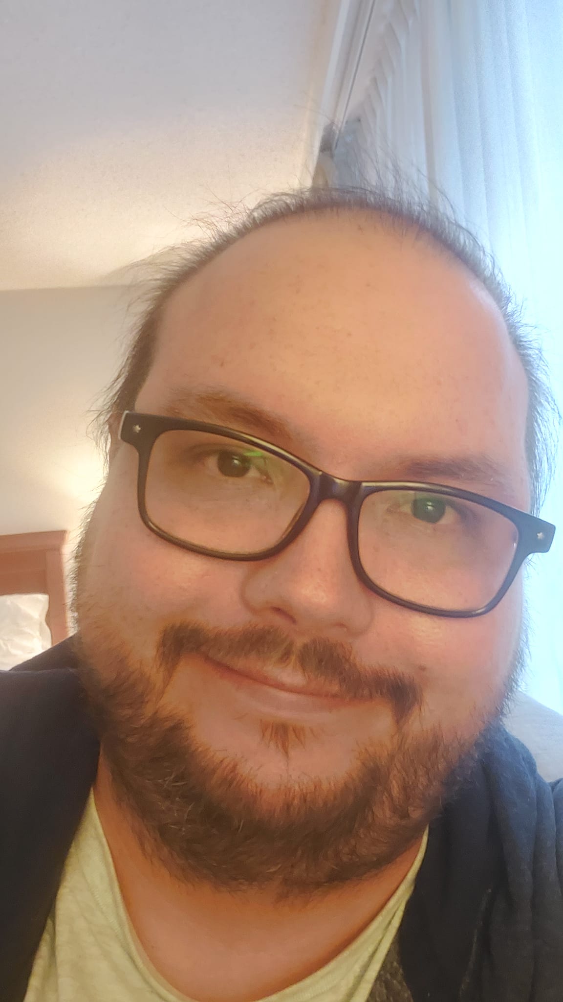 A man sits in a hotel room, smiling, with short fine hair and short facial hair